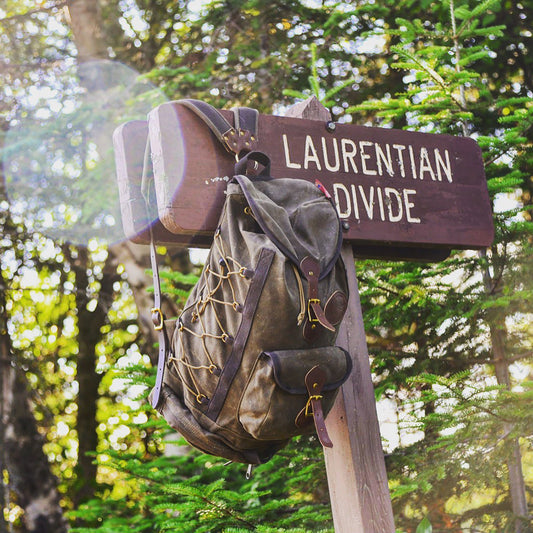 A vintage-style canvas backpack with brown leather straps and gold-toned hardware hangs from a wooden trail sign that reads 'LAURENTIAN DIVIDE' in a sunlit forest setting.