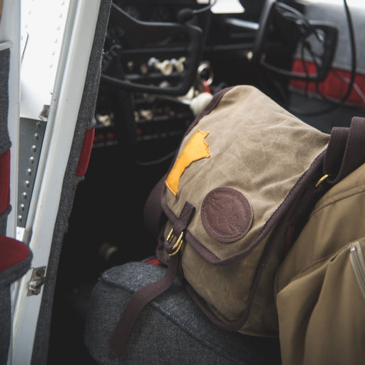 A smaller waxed canvas messenger bag sitting on a plane seat.