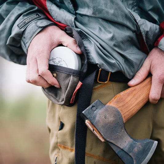 A man taking an axe sharpening puck out of his waxed canvas belt pouch. 