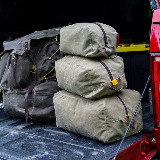 Three sizes of waxed canvas travel organizer bags stacked up and sitting on a tailgate at a campsite. 