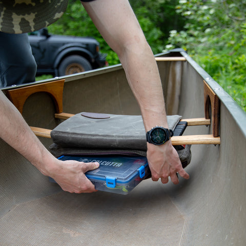 A person putting his box of fishing lures into the bag of Frost River's waxed canvas Canoe Seat Pad Bag. 