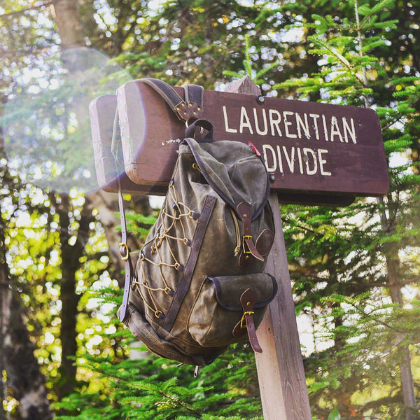 A vintage-style canvas backpack with brown leather straps and gold-toned hardware hangs from a wooden trail sign that reads 'LAURENTIAN DIVIDE' in a sunlit forest setting.