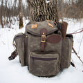 A rugged waxed canvas backpack in dark brown and olive tones, styled outdoors in the snow next to a tree trunk, with a brown leather sheath attached to the side.