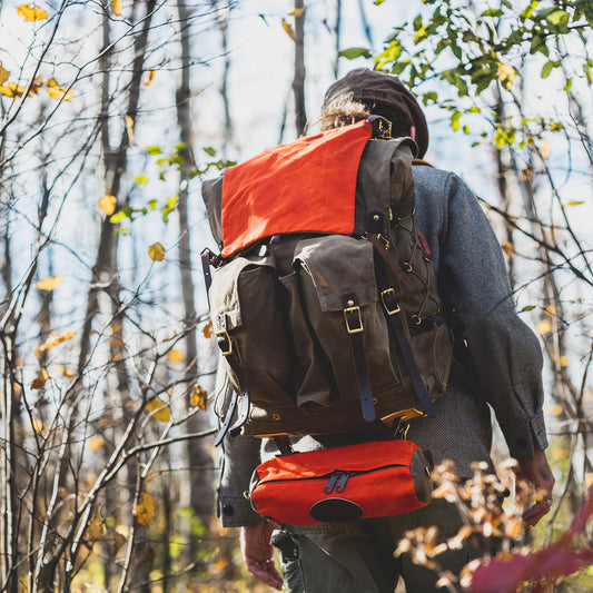 A person hiking through a wooded area seen from behind, wearing a large brown  backpack with a bright orange signal bag attatched.
