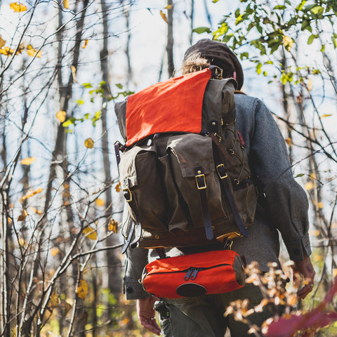 A person hiking through a wooded area seen from behind, wearing a large brown  backpack with a bright orange signal bag attatched.