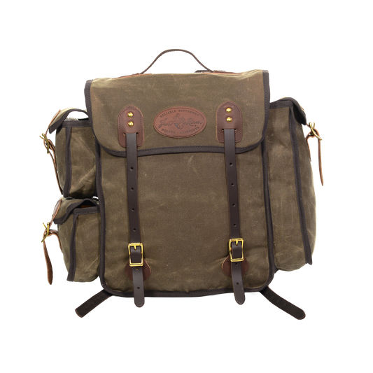A waxed canvas backpack with dark brown leather straps and trim, featuring a central flap pocket with brass-tone buckles and a circular leather logo patch.