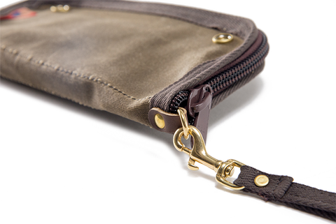 A small webbed cotton wrist strap is attached to the North Shore Clutch by a durable brass clip hook and brass attachment point.